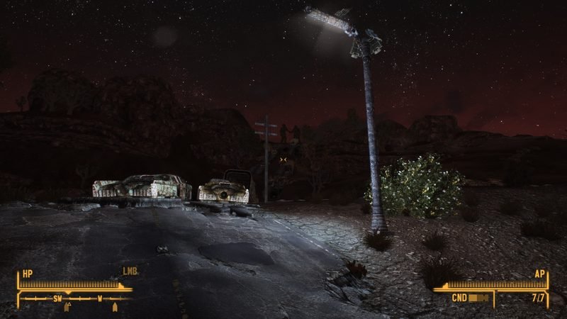 A working sodium street lamp lighting cracked asphalt and two wrecked cars at night, with figures on a ridge behind